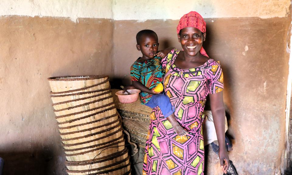A woman in colorful clothing holds a child in her arms, standing next to woven baskets against a textured wall.