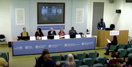 Photo of a panel of speakers seated behind a table beneath a large screen.