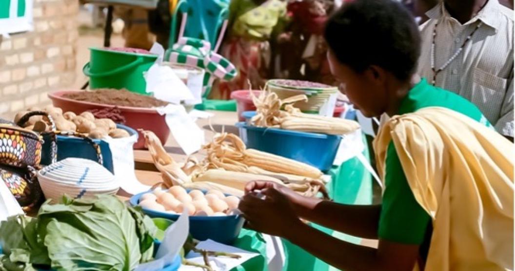 A woman in a yellow top and green apron stands at a market stall with various vegetables, eggs, and grains for sale.