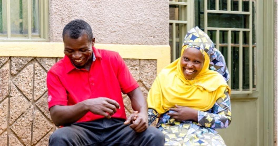Two individuals sit on a bench outside a building with yellow walls and windows with metal grills. They are both laughing and holding hands.