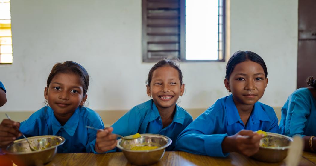 Three young children wearing blue school uniforms eat from bowls of freshly cooked vegetables.