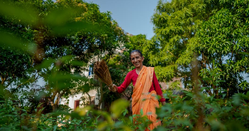 The image shows an elderly woman standing outdoors, smiling warmly while holding a broom made of natural materials. She is dressed in a vibrant orange sari with a red blouse, surrounded by lush greenery and trees.