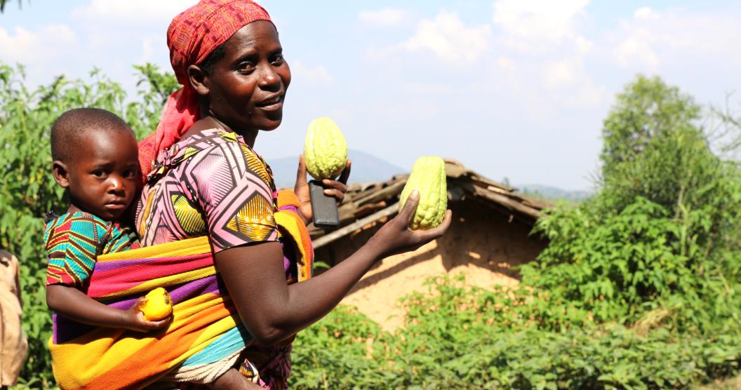 This image features a woman wearing colorful traditional clothing and carrying a child on her back in a vibrant, multi-colored wrap. She holds two bright green fruits (possibly gourds or similar produce) in her hands, while the child holds a yellow fruit.