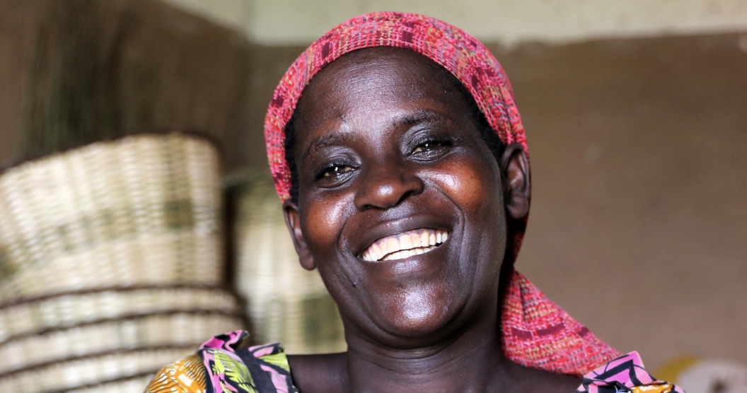 This image shows a smiling woman in vibrant, patterned clothing and a matching headscarf. Behind her, woven baskets are stacked, suggesting a setting related to agriculture, storage, or trade.